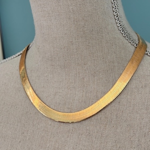 May include: A gold-colored herringbone necklace displayed on a mannequin. The necklace has a flat, wide design with a subtle, textured pattern. The necklace is a warm gold tone and is displayed against a neutral background.