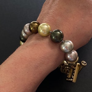May include: A bracelet made of large, round pearls in shades of green, gold, and grey. The bracelet has a gold clasp with a small black gemstone.
