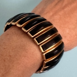Puede incluir: Una pulsera negra y dorada con un diseño acanalado. La pulsera está hecha de esmalte negro y metal dorado. La pulsera está en una muñeca.