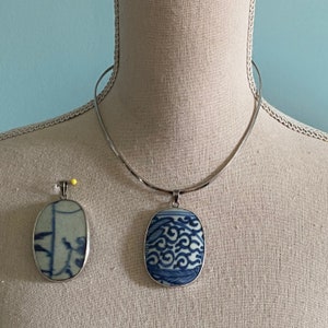 May include: A silver necklace with a blue and white porcelain pendant. The pendant is oval shaped and features a swirling pattern. The necklace is on a white fabric background.