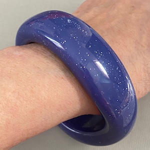May include: A wide, blue bangle bracelet with a sparkly finish. The bracelet is made of a smooth, glossy material and has a slightly rounded shape.