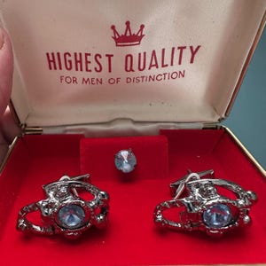 Könnte beinhalten: Ein Vintage-Manschettenknöpfe- und -Stecker-Set in einer rot samtgefütterten Box. Die silberfarbenen Manschettenknöpfe und der Stecker haben ein dekoratives Design mit einem großen, hellblauen Kristall in der Mitte. Der Deckel der Box lautet "HIGHEST QUALITY FOR MEN OF DISTINCTION".