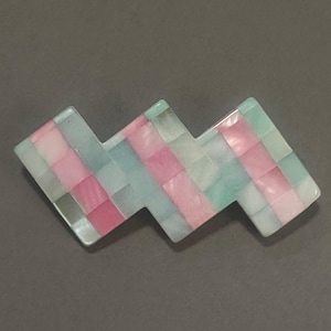 Puede incluir: Una pinza para el pelo con diseño de mosaico en forma de zigzag. La pinza presenta un patrón de pequeñas baldosas cuadradas en tonos rosa, azul y blanco. La pinza está hecha de un material brillante y está sobre un fondo gris.