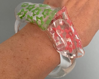 May include: A clear plastic bangle bracelet with a floral design. The bracelet has a green and red floral pattern.