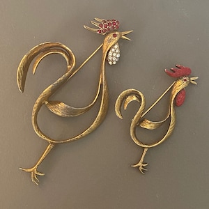 May include: Two gold-tone rooster brooches with red and clear rhinestone accents. The larger rooster has a more detailed design with a curved tail and a raised head. The smaller rooster has a simpler design with a straight tail and a flat head.