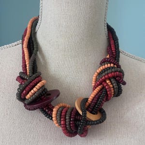 Può includere: Una collana a più fili con perline di legno nei toni del rosso, nero, grigio e pesca. La collana è annodata e presenta due grandi elementi rotondi in legno. La collana è esposta su un manichino.
