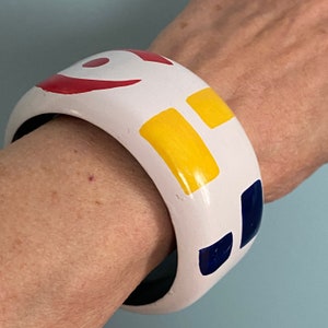 Op de afbeelding: Witte bangle armband met rode, gele en blauwe geometrische vormen erop geschilderd.
