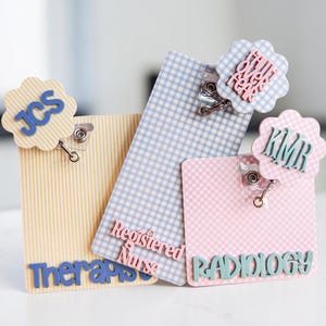Puede incluir: Tres portatarjetas rectangulares con clips met&aacute;licos y adornos festoneados. Uno amarillo a rayas con JCS, uno azul a cuadros con Registered Nurse y uno rosa a cuadros con RADIOLOGY y KMR. Un adorno incluye el texto Faith Over Fear.