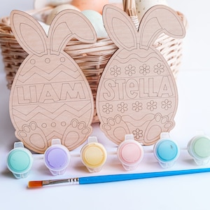 May include: Two unfinished wooden Easter bunny egg shapes with the names Liam and Stella painted on them. The eggs are in a wicker basket with other eggs. There are six small paint pots and a paintbrush on a white surface.