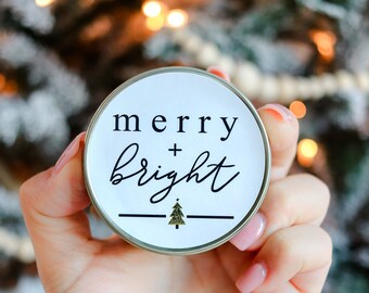 Puede incluir: Un recipiente redondo y blanco con las palabras "merry + bright" en escritura negra y un pequeño gráfico de árbol de Navidad dorado. El recipiente se sostiene en una mano, con un árbol de Navidad borroso en el fondo.