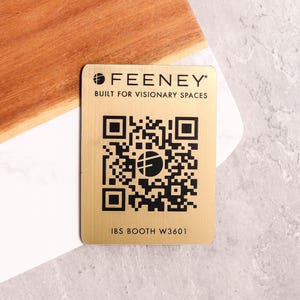 May include: A gold-colored metal card with a QR code and the text "FEENEY BUILT FOR VISIONARY SPACES." The card also includes the text "IBS BOOTH W3601." The card is on a white and gray surface with a wooden cutting board.