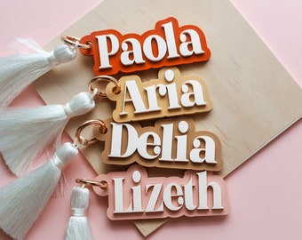 May include: Four personalized keychains with white tassels. Each keychain features a different name: Paola, Aria, Delia, and Lizeth. The names are in white, layered over colorful backgrounds of orange, tan, brown, and pink.