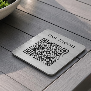 May include: A square, brushed metal sign featuring a black QR code and the text "our menu" printed in a simple sans-serif font. The sign is positioned on a slatted gray surface next to a white bowl filled with a green salad and a glass of water.