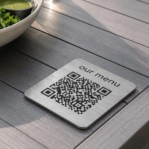 May include: A square, brushed metal sign featuring a black QR code and the text "our menu" printed in a simple sans-serif font. The sign has rounded corners and a flat, metallic finish, positioned on a slatted surface next to a bowl of salad.