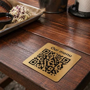 May include: A square gold-toned metal coaster featuring a black QR code and the text "Our menu" printed in black. The coaster has rounded corners and a smooth, reflective surface, positioned flat on a dark brown wooden table surface.