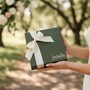 May include: A dark green gift box tied with a cream-colored ribbon. The box has the name "Samantha" written on it in a delicate script. The box is held by a person in a white dress.
