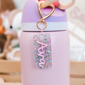 May include: A pink water bottle with a gold carabiner clip and a glittery acrylic tag that says "Ava" in pink script. The bottle has a purple lid and a pink body. The tag is filled with colorful glitter.