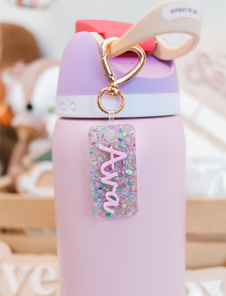 May include: A pink water bottle with a gold carabiner clip and a glittery acrylic tag that says "Ava" in pink script. The bottle has a purple lid and a pink body. The tag is filled with colorful glitter.