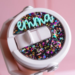 May include: White water bottle lid with the name "emma" in light blue script. The lid features a section filled with colorful, iridescent confetti against a black background. The bottle is held against a pink background.