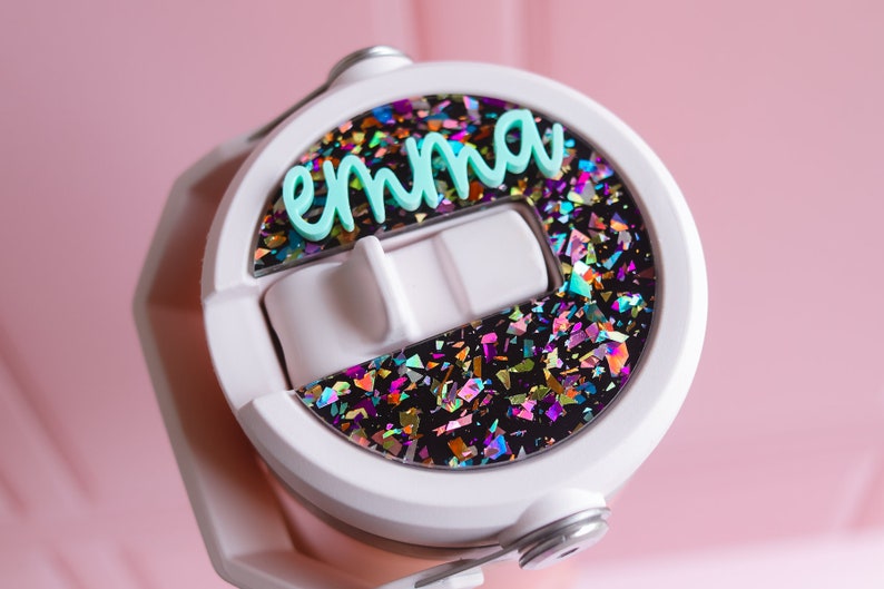 May include: White water bottle lid with the name "emma" in light blue script. The lid features a section filled with colorful, iridescent confetti against a black background. The bottle is held against a pink background.