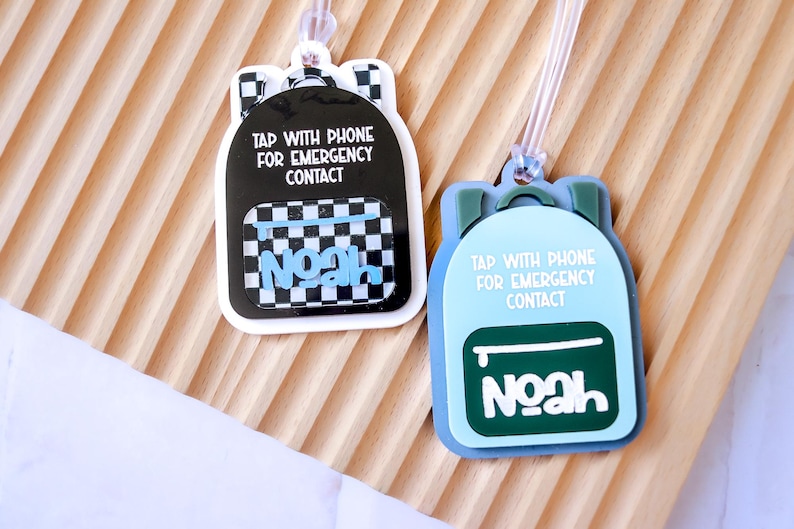 May include: Two backpack-shaped luggage tags. One is black with a checkered pocket and the name "Noah" in blue. The other is light blue with a green pocket and the name "Noah" in white. Both tags have the text "TAP WITH PHONE FOR EMERGENCY CONTACT".