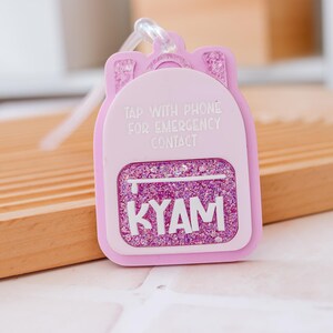 Puede incluir: Un llavero rosa con forma de mochila con detalles de purpurina y el nombre "KYAM" en blanco. El llavero tiene el texto "TAP WITH PHONE FOR EMERGENCY CONTACT".