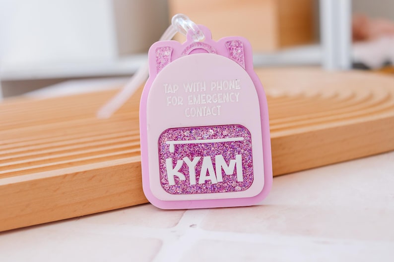Puede incluir: Un llavero rosa con forma de mochila con detalles de purpurina y el nombre "KYAM" en blanco. El llavero tiene el texto "TAP WITH PHONE FOR EMERGENCY CONTACT".