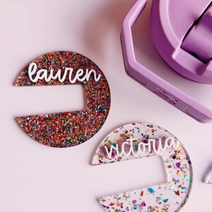 May include: Two half-circle shaped name tags with the names "Lauren" and "Victoria" in white script. The "Lauren" tag is covered in colorful glitter, while the "Victoria" tag has confetti. A purple water bottle is in the background.