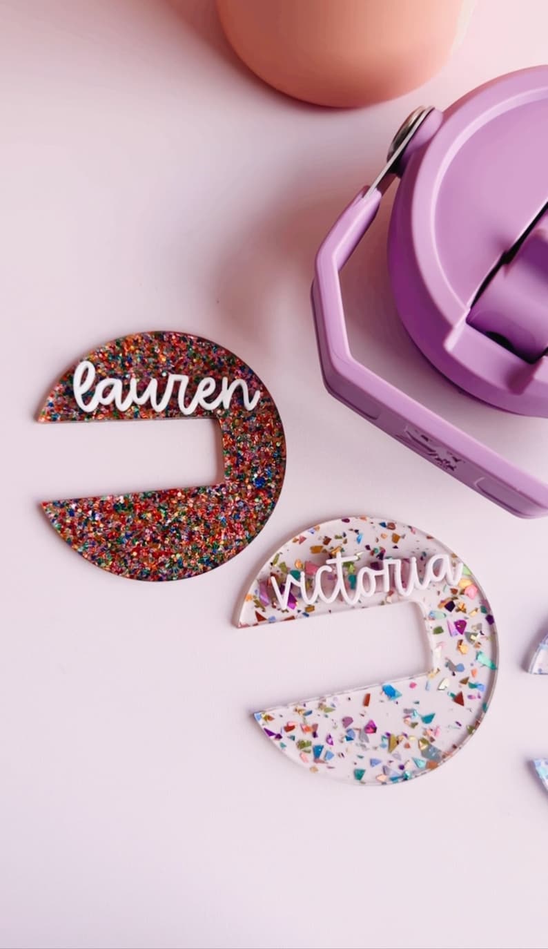 May include: Two half-circle shaped name tags with the names "Lauren" and "Victoria" in white script. The "Lauren" tag is covered in colorful glitter, while the "Victoria" tag has confetti. A purple water bottle is in the background.