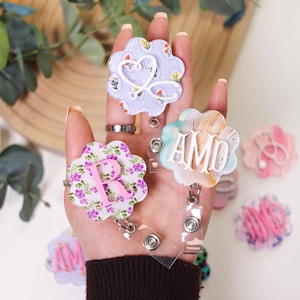 May include: A selection of flower-shaped badge reels, each with a personalized initial. The reels display diverse designs, such as floral patterns, watercolour effects, and pastel shades. Each reel has a retractable cord and a clear plastic clip.