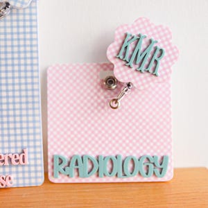 Puede incluir: Dos portatarjetas rectangulares con patrones de cuadros vichy. El de la izquierda es azul claro con texto rosa que dice Registered Nurse. El de la derecha es rosa claro con texto verde azulado RADIOLOGY y un adorno festoneado con las iniciales KMR.
