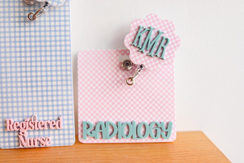 Puede incluir: Dos portatarjetas rectangulares con patrones de cuadros vichy. El de la izquierda es azul claro con texto rosa que dice Registered Nurse. El de la derecha es rosa claro con texto verde azulado RADIOLOGY y un adorno festoneado con las iniciales KMR.