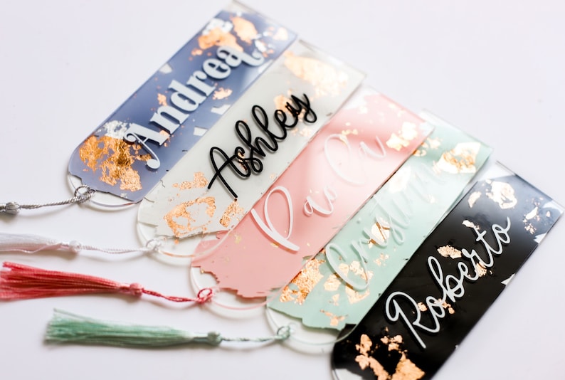 May include: Five personalized bookmarks with gold leaf accents and tassels. The bookmarks are made of clear acrylic and feature the names Andrea, Ashley, Paula, Christina, and Roberto.