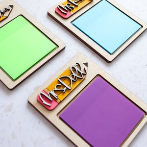 Puede incluir: Tres porta notas de madera con un diseño de lápiz y el texto "Mrs. Saliba", "Mrs. Wish" y "Mrs. Dell". Cada porta notas tiene un espacio cuadrado para una nota adhesiva. Las notas adhesivas son verde, azul y morado.