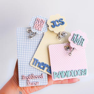 Puede incluir: Tres portatarjetas rectangulares con patrones de cuadros vichy en azul, amarillo y rosa. Cada uno tiene un adorno festoneado con textos como "faith over fear", "JCS" y "KMR". El azul dice "Registered Nurse", el amarillo "Therapy" y el rosa "RADIOLOGY".