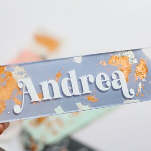 May include: A clear rectangular name tag with the name "Andrea" in white lettering. The tag has a light blue background with gold and silver flecks.