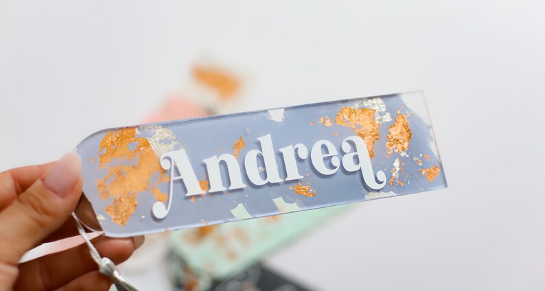 May include: A clear rectangular name tag with the name "Andrea" in white lettering. The tag has a light blue background with gold and silver flecks.