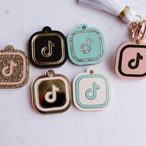 May include: Five different keychains with a TikTok logo. The keychains are square and have a black, gold, pink, blue, and white background. The TikTok logo is in black, gold, pink, blue, and white on each keychain.