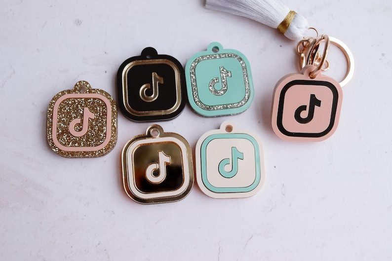 May include: Five different keychains with a TikTok logo. The keychains are square and have a black, gold, pink, blue, and white background. The TikTok logo is in black, gold, pink, blue, and white on each keychain.