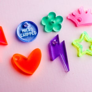 Pu&ograve; includere: Assortimento di ciondoli in acrilico colorati in varie forme, tra cui un cuore, un fulmine, una stella, un fiore, una corona e un ciondolo rotondo con il testo "MICRO CHIPPED".