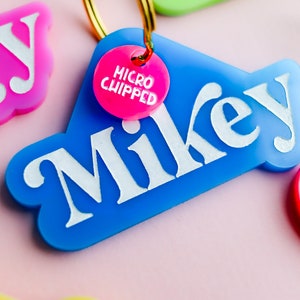 Pu&ograve; includere: Un'etichetta per il nome in plastica blu con la parola "Mikey" in lettere bianche. Un'etichetta in plastica rosa con le parole "MICRO CHIPPED" &egrave; attaccata all'etichetta blu con un anello dorato.