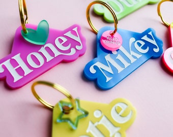 May include: Four colorful plastic pet ID tags with gold key rings. The tags are shaped like a heart, a star, and two irregular shapes. The tags are labeled "Honey", "Mikey", "Otie", and "C". One tag has a pink circle with the text "Micro Chipped".