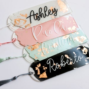 May include: Four personalized bookmarks with gold foil accents and tassels. The bookmarks are made of clear acrylic and feature the names "Ashley", "Paola", "Cristina", and "Roberto" written in black and white lettering.