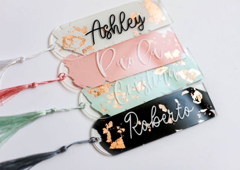 May include: Four personalized bookmarks with gold foil accents and tassels. The bookmarks are made of clear acrylic and feature the names "Ashley", "Paola", "Cristina", and "Roberto" written in black and white lettering.