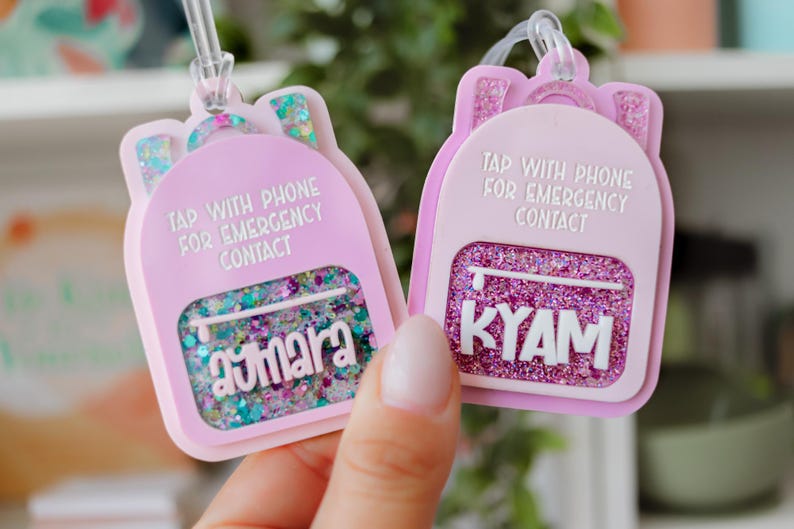 May include: Two pink backpack-shaped keychains with glitter and the text "Tap with phone for emergency contact." One keychain has the name "Aymara" and the other has "KYAM". Both have a clear loop for attachment.