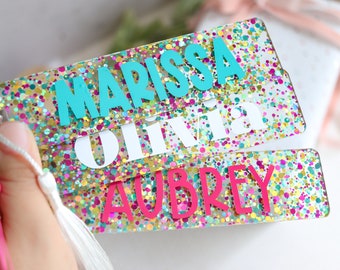 May include: Three colorful glitter bookmarks with the names "Marissa", "Olivia", and "Aubrey" written in different colors. The bookmarks have a white tassel.