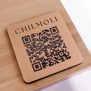 Peut inclure: Un dessous de verre en bois brun avec le texte "CHILMOLI" et un code QR noir et blanc.