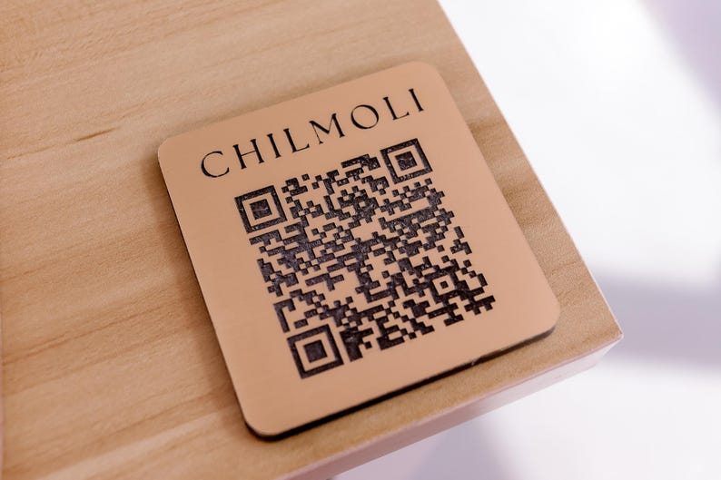 Peut inclure: Un dessous de verre en bois brun avec le texte "CHILMOLI" et un code QR noir et blanc.
