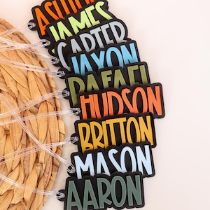 May include: A collection of colorful name tags with various names printed in bold, outlined letters. The tags are black with names in orange, green, blue, and other colors. Clear plastic loops are attached for hanging. The tags are arranged near a woven, circular placemat.