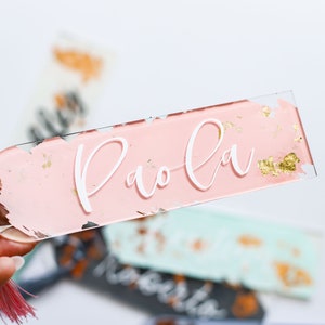 May include: A pink acrylic bookmark with gold flecks and the name "Paola" written in white lettering. The bookmark has a pink tassel.